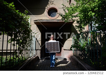 Woman holding solar panel for charging, stands in front of old building's entrance. Smiling girl wears sunglasses, white top, and blue jeans. Scene framed by green foliage and iron fencing. 120481512