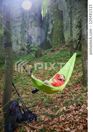 Person relaxing in bright green hammock tied between two trees in lush, green forest near rocks. Man tourist holds drink, enjoying peaceful moment surrounded by nature. Backpack and gear rest nearby. 120481515