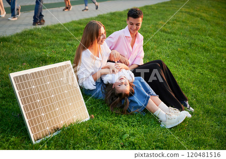 Young women using solar panel for charging, sit on grassy lawn, smiling and interacting with small child lying playfully on woman lap. Girls casually dressed, enjoying sunny, relaxed moment outdoors. 120481516
