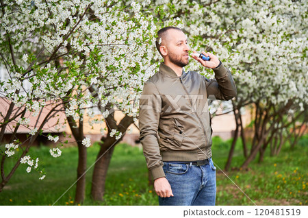 Man allergic using medical nasal drops, suffering from seasonal allergy at spring in blossoming garden. Handsome man treating runny nose in front of blooming tree outdoors. Spring allergy concept. 120481519