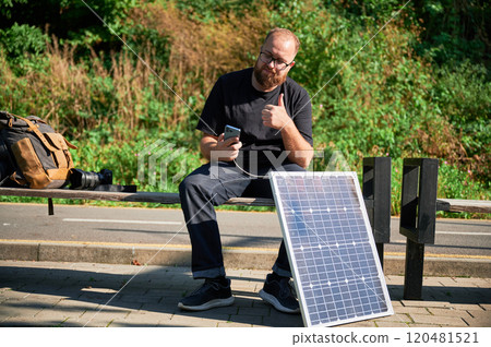 Bearded man using smartphone connected to photovoltaic solar panel, giving thumbs up. Integration of sustainable renewable energy into everyday life, practical use of solar power for charging devices. Bearded man using smartphone connected to photovoltaic solar panel, giving thumbs up. Integration of sustainable renewable energy into everyday life, practical use of solar power for charging devices. 120481521