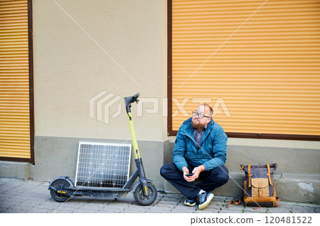 Male person waits while charging electric scooter with photovoltaic solar panel in urban settings. Integration of solar power as sustainable energy source, promoting eco-friendly urban transportation. 120481522