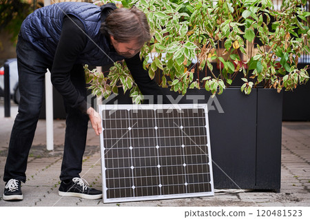 Man crouches down to place solar panel near large planter with green foliage. Guy in black outfit and navy blue quilted vest. Concept on integrating sustainable energy with city life. Man crouches down to place solar panel near large planter with green foliage. Guy in black outfit and navy blue quilted vest. Concept on integrating sustainable energy with city life. 120481523