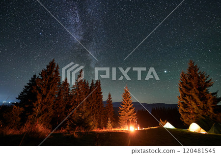 Panoramic view of mountain hill with illuminated camp tents, coniferous trees and hikers under starry sky. Night camping in mountains under beautiful sky with stars. Concept of hiking and traveling. 120481545