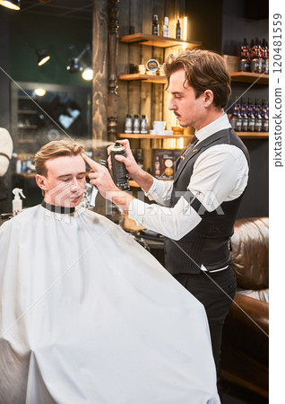 Professional barber using hairspray to finish hairstyle. Stylish hairdresser making final touches. Young customer getting fresh haircut. Professional barber using hairspray to finish hairstyle. Stylish hairdresser making final touches. Young customer getting fresh haircut. 120481559
