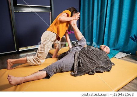 Masseuse making thai yoga massage. Woman therapist doing traditional massage treatment, stretching male patient's body. Bald client lying on mat, enjoying hand massage in calm atmosphere. Masseuse making thai yoga massage. Woman therapist doing traditional massage treatment, stretching male patient's body. Bald client lying on mat, enjoying hand massage in calm atmosphere. 120481568