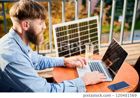 Man works on laptop and smartphone at outdoor table, with glass of iced coffee. Solar panel nearby, emphasizing sustainable, modern eco-friendly workspace that combines technology and sustainability. Man works on laptop and smartphone at outdoor table, with glass of iced coffee. Solar panel nearby, emphasizing sustainable, modern eco-friendly workspace that combines technology and sustainability. 120481569