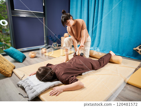 Masseuse making thai yoga massage. Woman therapist doing traditional massage treatment, stretching female patient's body. Client lying on mat, enjoying hand massage in calm atmosphere. 120481572