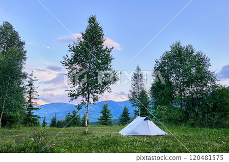 White tourist tent on grassy clearing, surrounded by lush trees with distant blue mountains under sky tinged with pink clouds. Peaceful scene exudes tranquility of nature and serene mountain camping. 120481575
