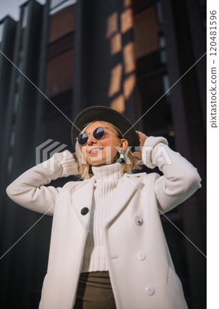 Stylish woman in a white coat and hat poses confidently outdoors against a modern urban backdrop Stylish woman in a white coat and hat poses confidently outdoors against a modern urban backdrop 120481596