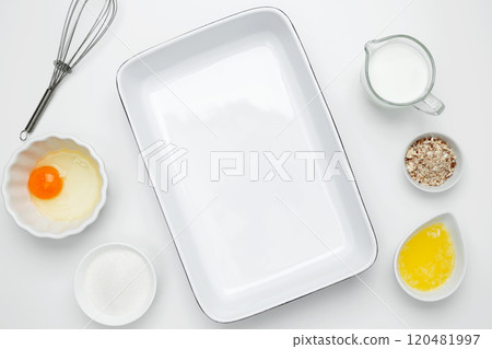 Empty white baking dish with ingredients for baking, including egg, milk, butter, sugar, and nuts on a white background. Flat lay composition for cooking preparation 120481997