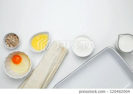 Near baking dish with ingredients, phyllo dough, egg, butter, nuts, and milk on a white background for baking Crinkle Cake 120482004