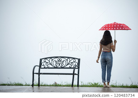 Woman holding red polka dot umbrella standing near metal bench on rainy day with overcast sky Woman holding red polka dot umbrella standing near metal bench on rainy day with overcast sky 120483734