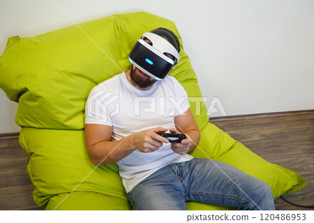 Man leaning back on green bean bag, wearing VR headset, and holding gaming controller 120486953