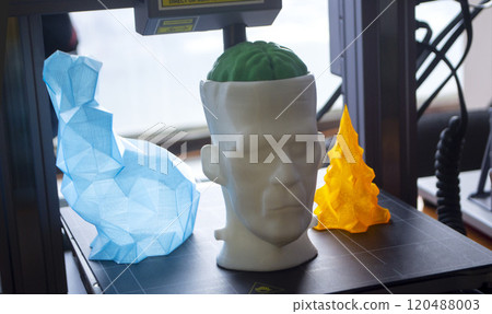 Art models printed on 3D printer on desktop of 3D printer. Models of prototype human head with brain, figured hare and Christmas tree printed on 3D printer from molten plastic of different colors 120488003