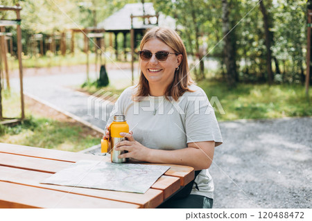 Happy 30s Woman in active trekking clothes having a halt after hiking. Hiker drinking water from water bottle or hot drink from yellow thermos. Woman planning new travel with paper map 120488472