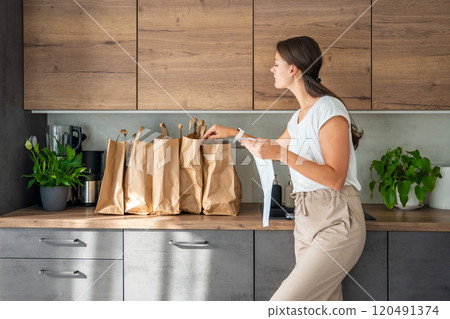 Young woman with receipt from the store next to paper bags full of food in kitchen. Groceries ordered online and delivered by courier 120491374
