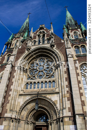 A close-up view of a neo-Gothic cathedral facade featuring intricate stone carvings, pointed arches, and a large rose window, set against a bright blue sky. 120491824