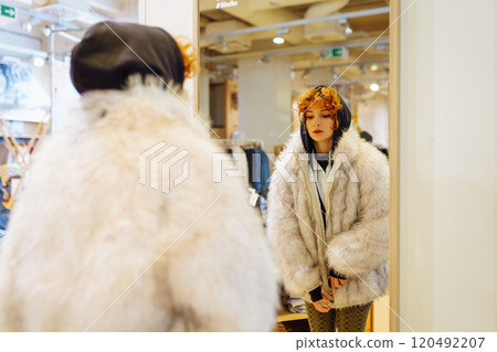 portrait of red-haired attractive young woman trying on fashionable fur coat in store 120492207
