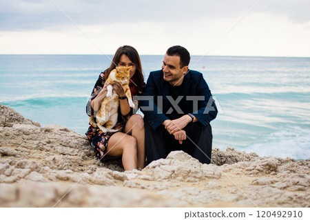 a young couple in love playing with orange stray cat on fabulous rocky coast with turquoise water Ayia Napa, Cyprus 120492910