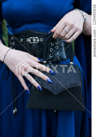 Close-up of a woman wearing a formal blue dress in a gottic style, her hands with an attractive manicure, holding a glittering handbag. Selective focus. Close-up of a woman wearing a formal blue dress in a gottic style, her hands with an attractive manicure, holding a glittering handbag. Selective focus. 120493916