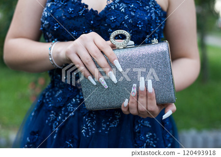 Close-up of a woman wearing a formal blue dress, her hands with an attractive manicure, holding a glittering handbag. Selective focus. Close-up of a woman wearing a formal blue dress, her hands with an attractive manicure, holding a glittering handbag. Selective focus. 120493918