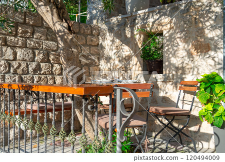 A charming courtyard with a wooden table and chairs, surrounded by stone walls and greenery, basking in sunlight and natural serenity 120494009