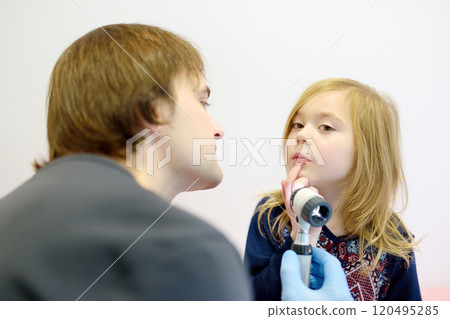 Cute little girl during appointment of dermatologist in modern clinic. Male doctor looking a pimple on a lips of child. Treatment of herpes virus, streptococcus infection 120495285