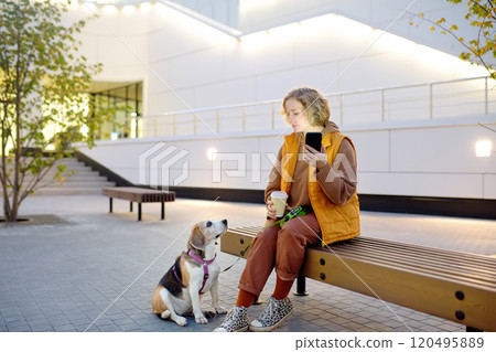 Hipster woman drinks coffee while walking with old Beagle dog along city street. Girl, dog owner, searches for pet products on the Internet using smartphone applications. Vet emergency care for pets 120495889