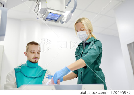 Young man having x-ray shot of broken hand in x ray room in modern clinic. Patient wearing in protect lead apron 120495982