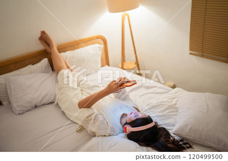 A woman relaxes in bed with her smartphone, enjoying a cozy atmosphere A woman relaxes in bed with her smartphone, enjoying a cozy atmosphere 120499500