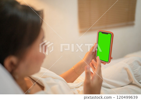 A woman relaxes in bed with a smartphone displaying a green screen, enjoying calm A woman relaxes in bed with a smartphone displaying a green screen, enjoying calm 120499639