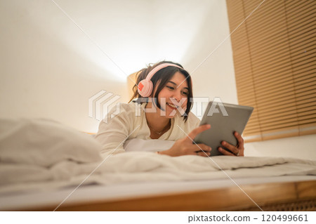Young Woman Relaxing and Enjoying Music on Her Tablet in a Cozy Bedroom Setting 120499661