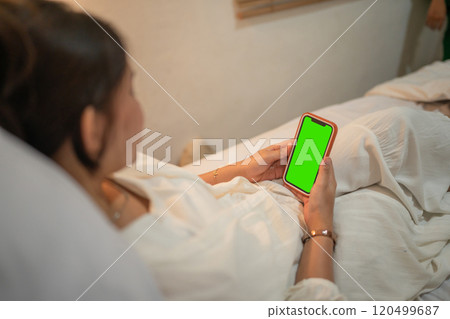 A woman relaxes in bed using a smartphone with a green screen in a cozy setting 120499687