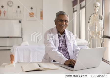 Mature Indian otolaryngologist posing for camera seated desk with laptop 120505836