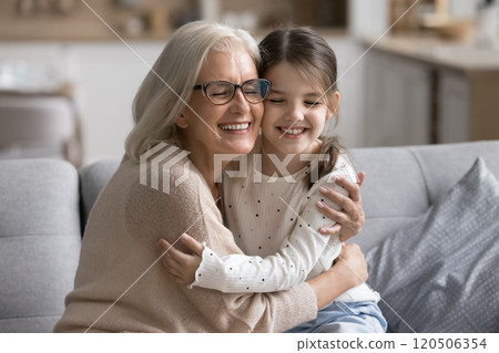 Heartwarming moment between grandmother and granddaughter embracing seated on sofa 120506354