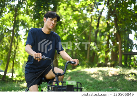 Young man riding a rental bike along a shaded dirt path surrounded by trees in a serene park 120506849