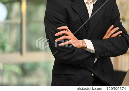 Confident businessman in sleek black suit standing with arms crossed in modern office 120506874