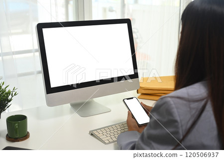 Side view of female employee holding smartphone with empty screen sitting in front of computer desktop Side view of female employee holding smartphone with empty screen sitting in front of computer desktop 120506937