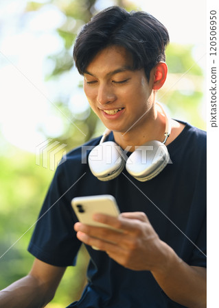 Smiling asian man with headphones around his neck using mobile phone in a park Smiling asian man with headphones around his neck using mobile phone in a park 120506950