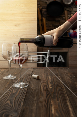 Pouring red wine into glasses on a rustic wooden table in a cozy kitchen at sunset Pouring red wine into glasses on a rustic wooden table in a cozy kitchen at sunset 120507779