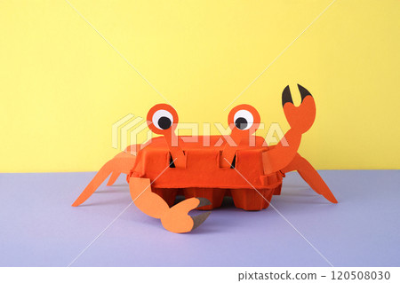 Recycled crab craft using red egg carton with paper details for legs, claws, and eyes. Waving claw and smiling face on purple background with yellow surface. Fun eco-friendly DIY idea, 120508030