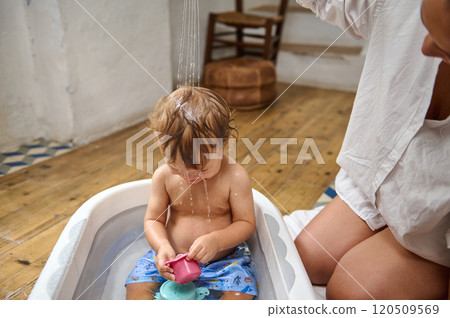 A cute toddler sits in a small bathtub, playfully enjoying water streaming down their head, creating a joyful and carefree atmosphere in a cozy room. A cute toddler sits in a small bathtub, playfully enjoying water streaming down their head, creating a joyful and carefree atmosphere in a cozy room. 120509569