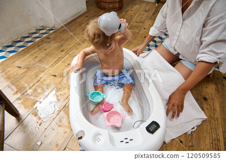 A joyful toddler enjoys a bath, playing with water toys, while a caring parent ensures safety and fun. The scene captures warmth and parental love on a wooden floored interior. A joyful toddler enjoys a bath, playing with water toys, while a caring parent ensures safety and fun. The scene captures warmth and parental love on a wooden floored interior. 120509585