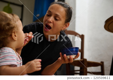 A mother lovingly feeds her baby with a spoon in a cozy kitchen setting, showcasing warmth and care. The child's curiosity and the mother's tenderness highlight a nurturing bond. 120509611