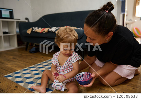 A mother lovingly assists her child in painting, creating cherished memories in a cozy home setting. The scene radiates warmth and creativity as they bond over a fun activity. 120509638