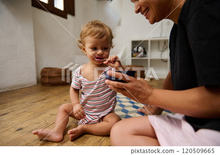 A mother feeds her adorable toddler with a smile in a cozy home interior. The child enjoys a meal in a warm, loving atmosphere, showcasing family bonding and joyful parenting moments. A mother feeds her adorable toddler with a smile in a cozy home interior. The child enjoys a meal in a warm, loving atmosphere, showcasing family bonding and joyful parenting moments. 120509665