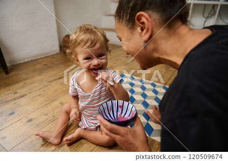 A delightful scene of a parent joyfully feeding their toddler in a cozy home setting. The child's bright smile and the colorful bowl of food create a warm, family-focused atmosphere. 120509674