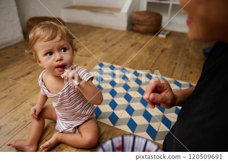 A charming toddler in a striped outfit sits on a wooden floor, eagerly tasting food from a spoon. The cozy indoor environment adds warmth and a sense of playful curiosity. 120509691