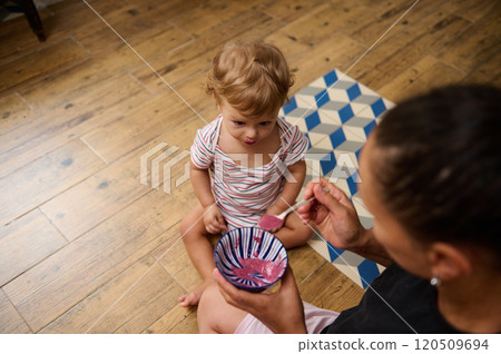 A mother lovingly feeds her curious toddler with a spoonful of pink yogurt while they sit on a wooden floor. A mother lovingly feeds her curious toddler with a spoonful of pink yogurt while they sit on a wooden floor. 120509694